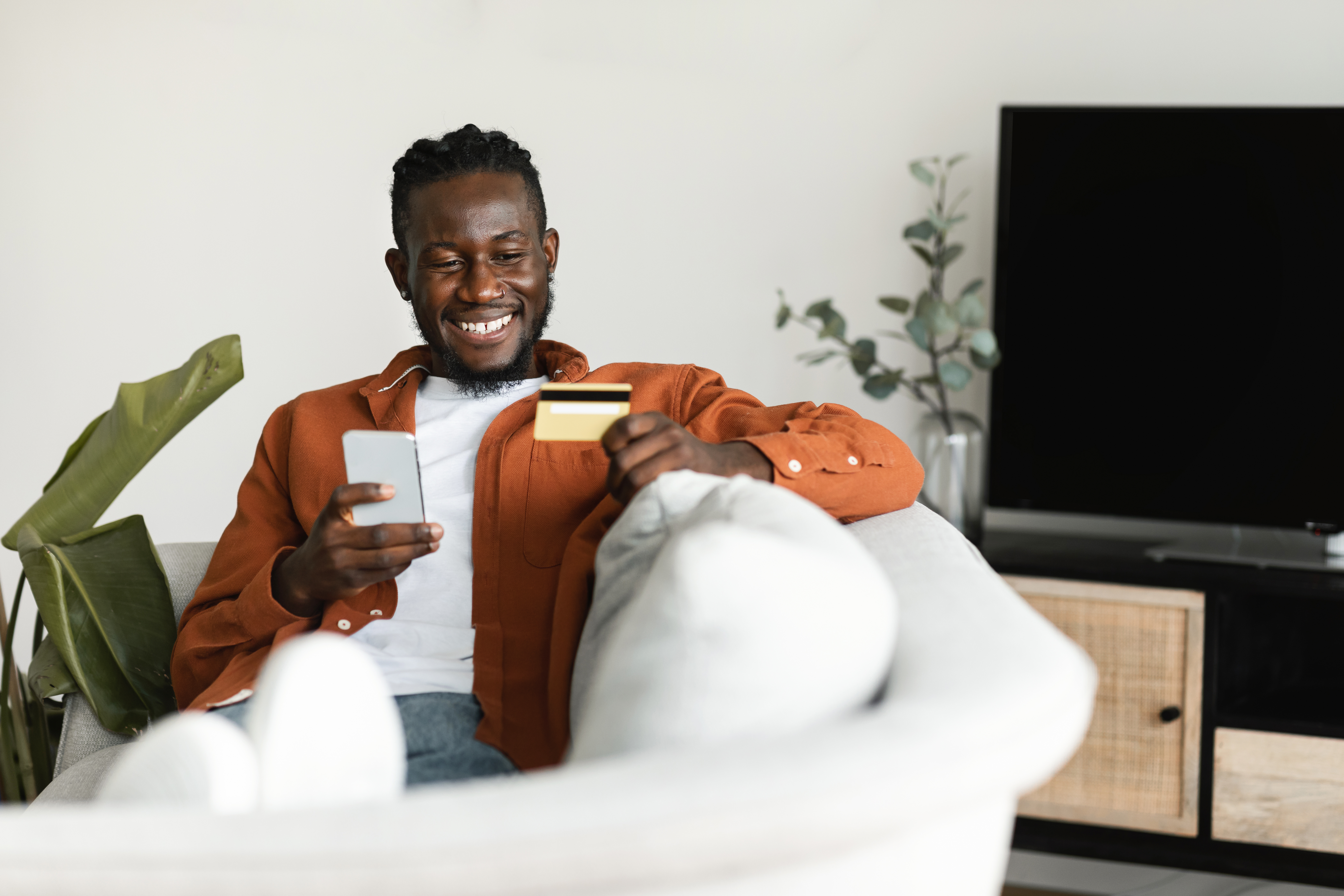 Man Holding Debit Credit Card in Hand and Using Smartphone, Making Financial Transaction While Resting on Couch at Home