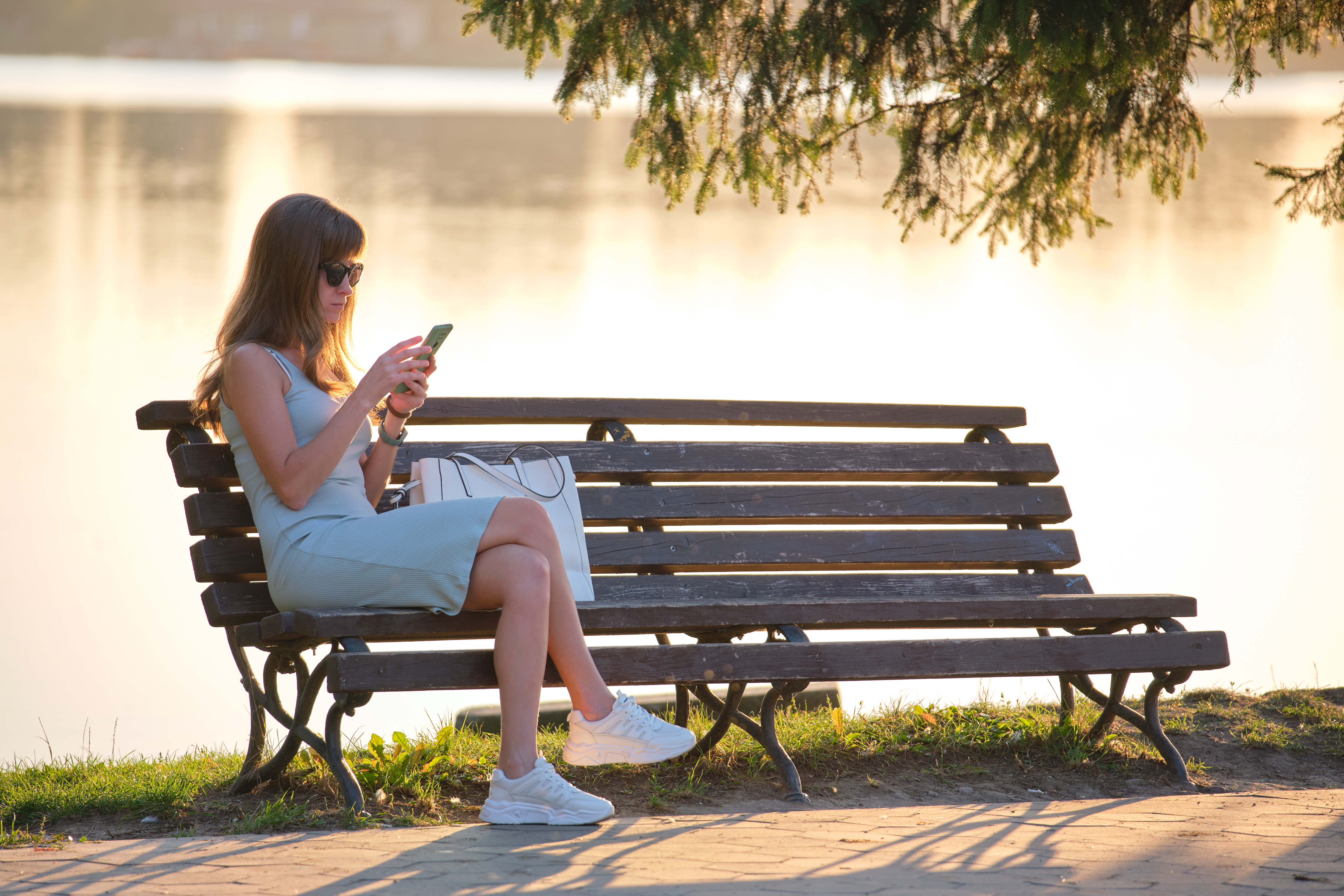 Young woman sitting on park bench browsing her cellphone outdoors on warm summer evening Young woman sitting on park bench browsing her cellphone outdoors on warm summer evening